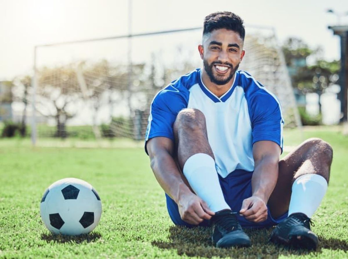 Young man lacing his boots in a field with football beside him