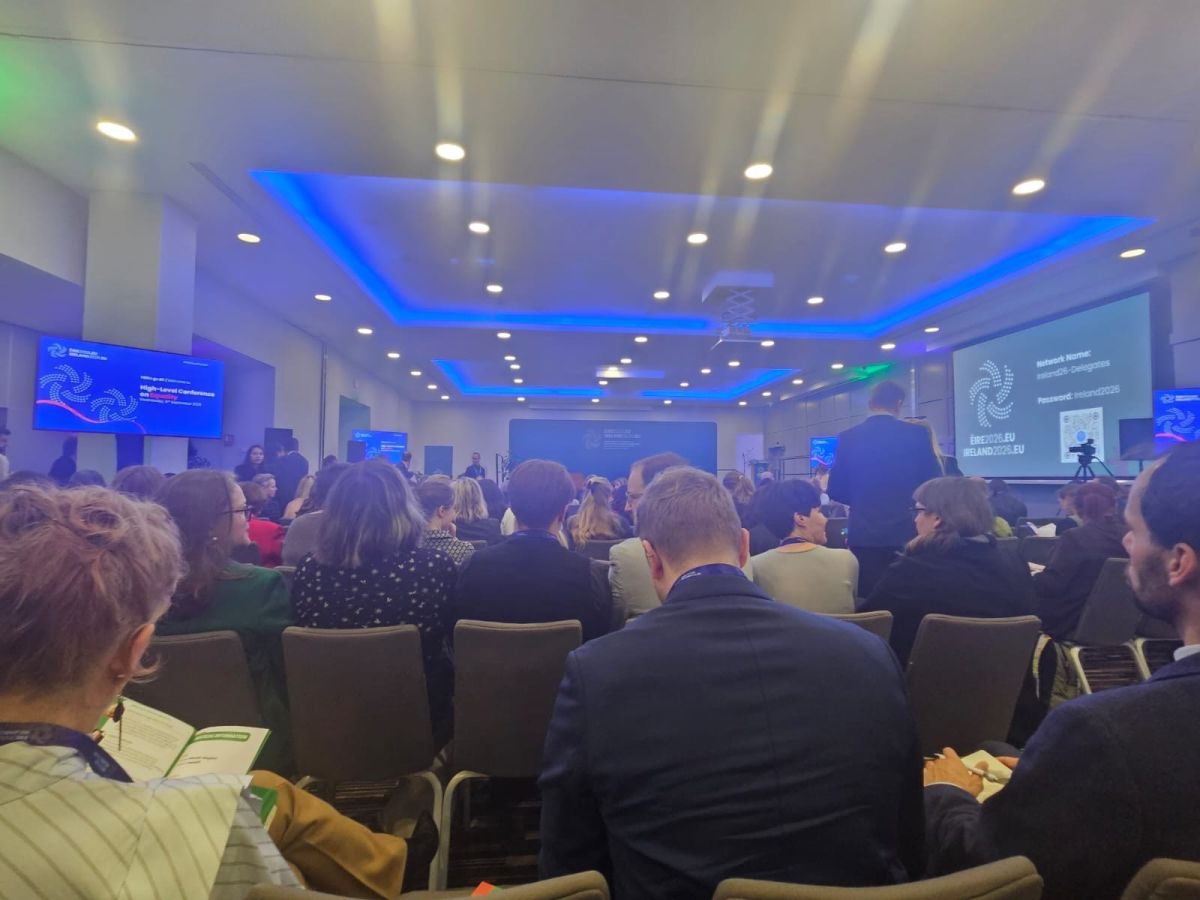 Conference audience seated in modern auditorium with blue ceiling lighting and multiple display screens showing presentations